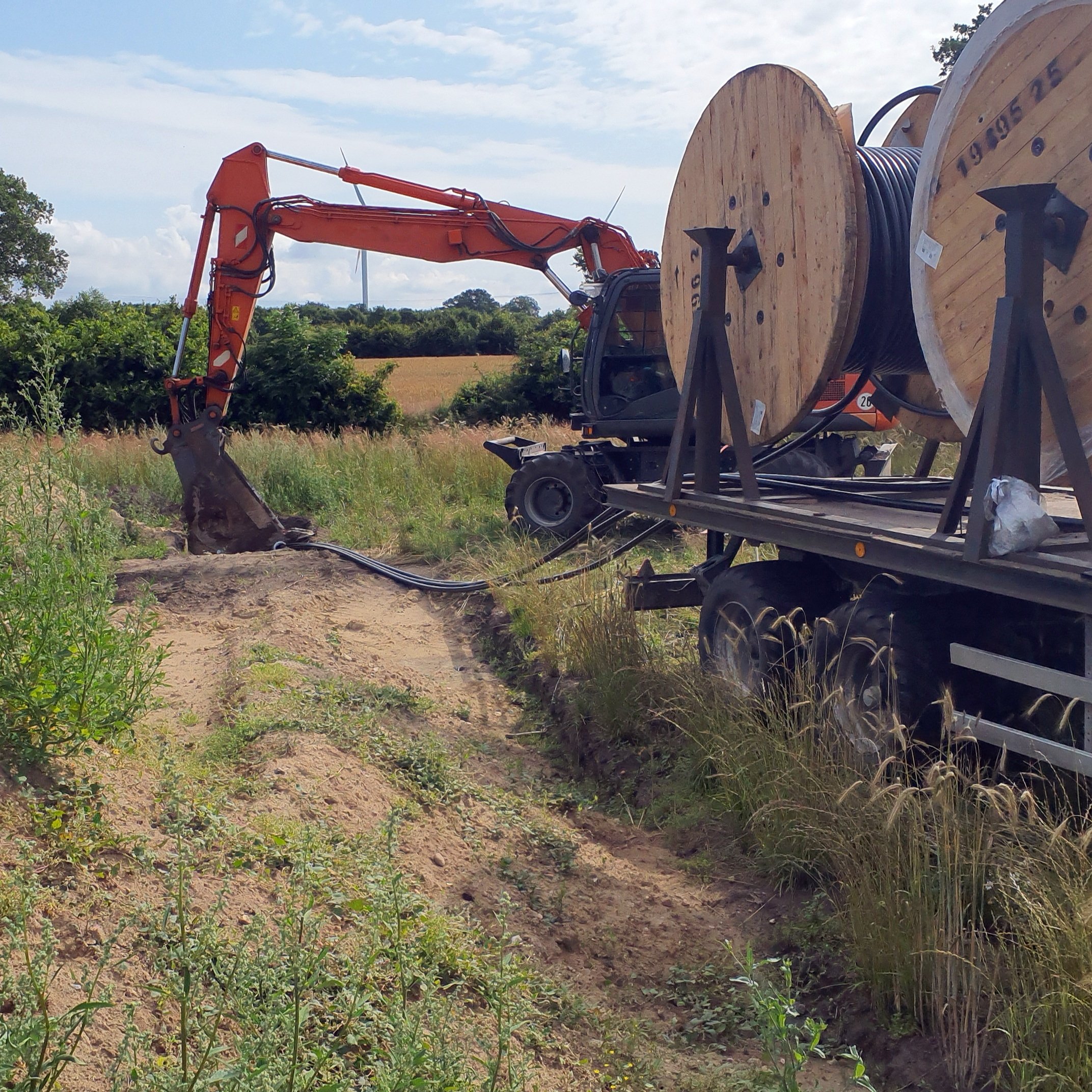 Ein Bagger steht hinter einem Anhänger mit Kabeltrommeln, auf denen Stromkabel aufgerollt sind. Im Hintergrund Büsche, Bäume und ein Feld. 