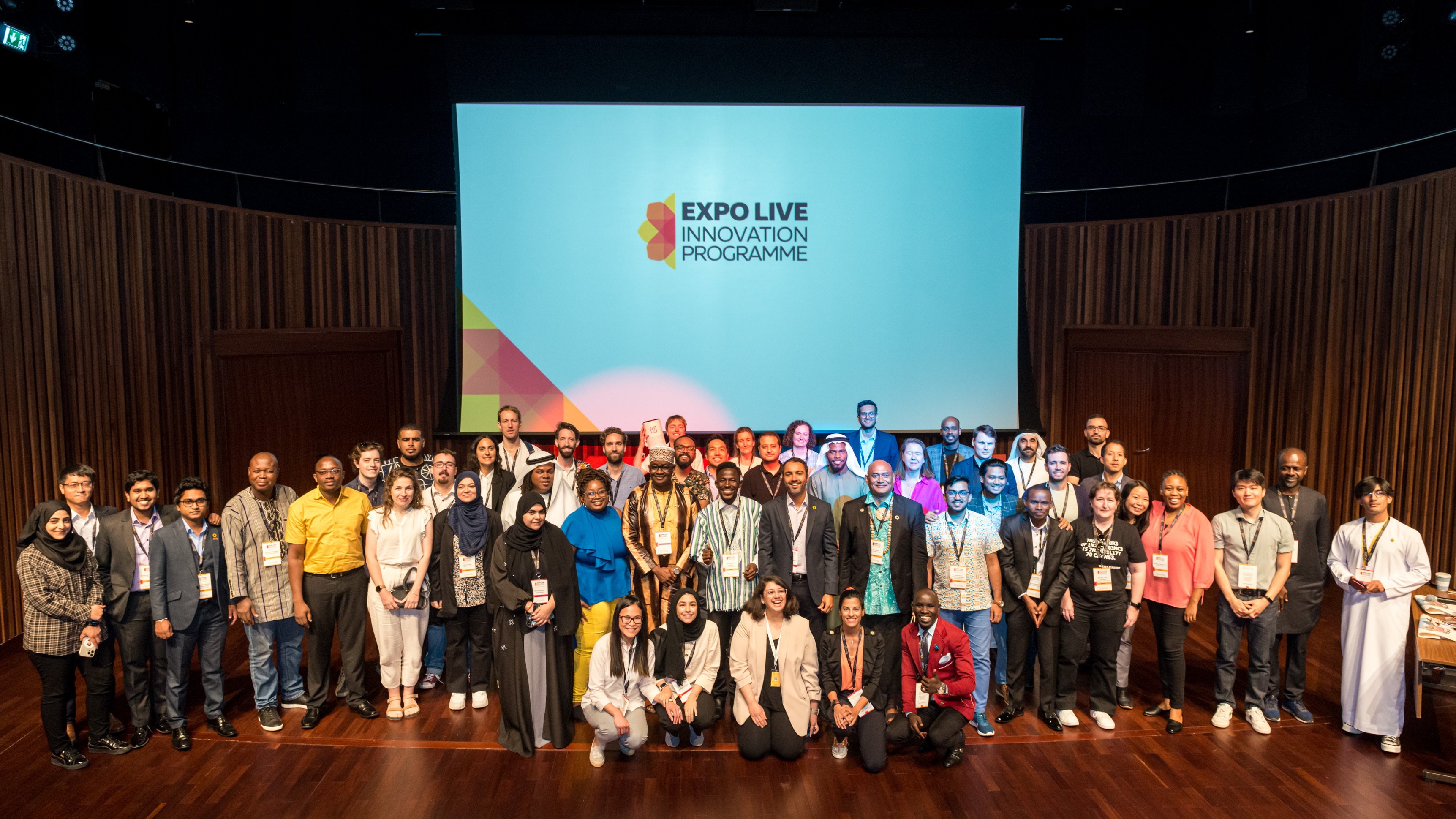 Ein Gruppenfoto, ca. 60 Menschen. Dahinter ein großer Bildschirm, auf dem das Logo des Expo Live Innovation Programms zu sehen ist.
