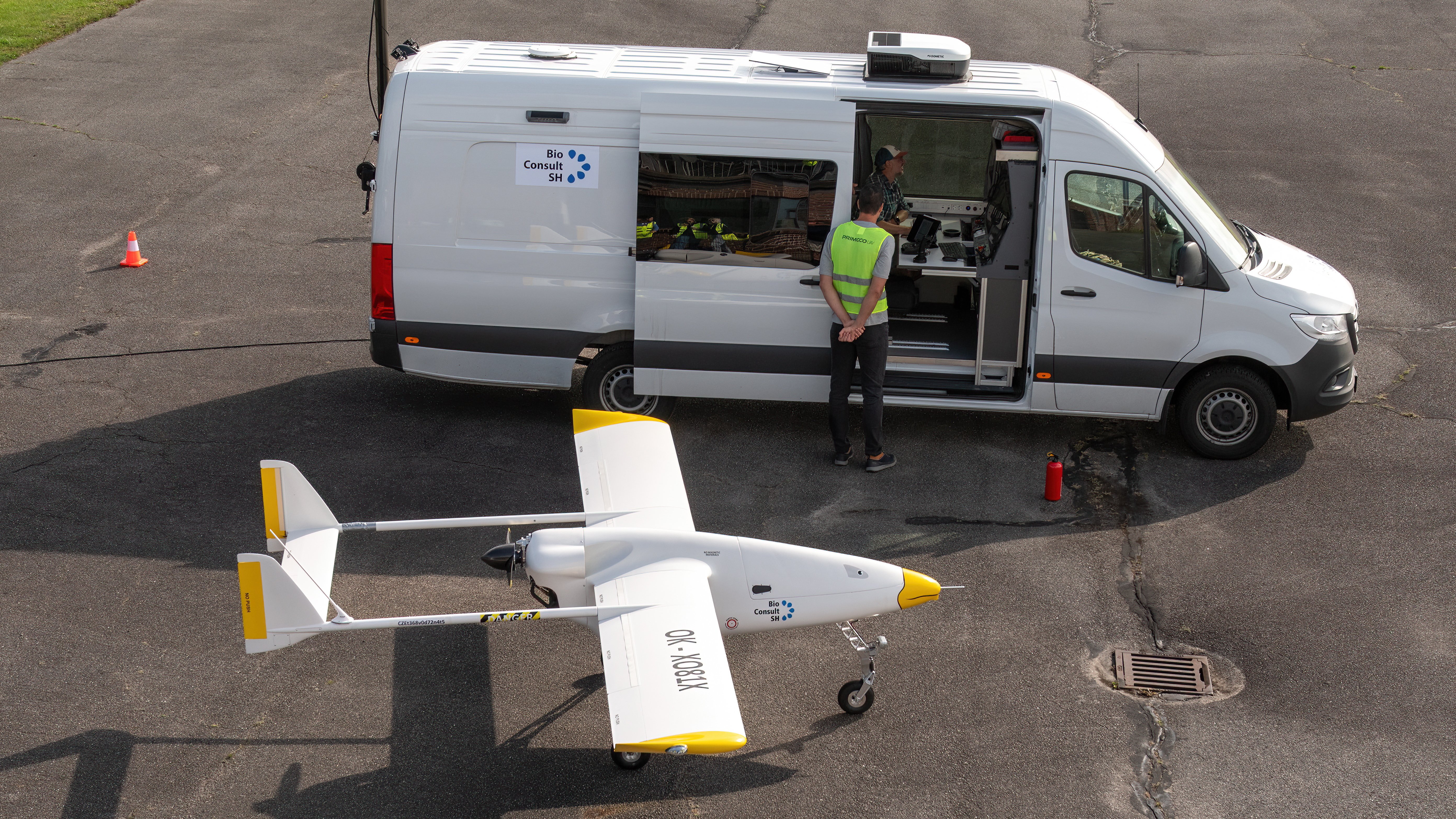 Die weiße Langstreckendrohne Primoco UAV One 150 steht auf dem Flugfeld, dahinter ein weißer Van, von dem aus die Drohne gesteuert und überwacht wird.