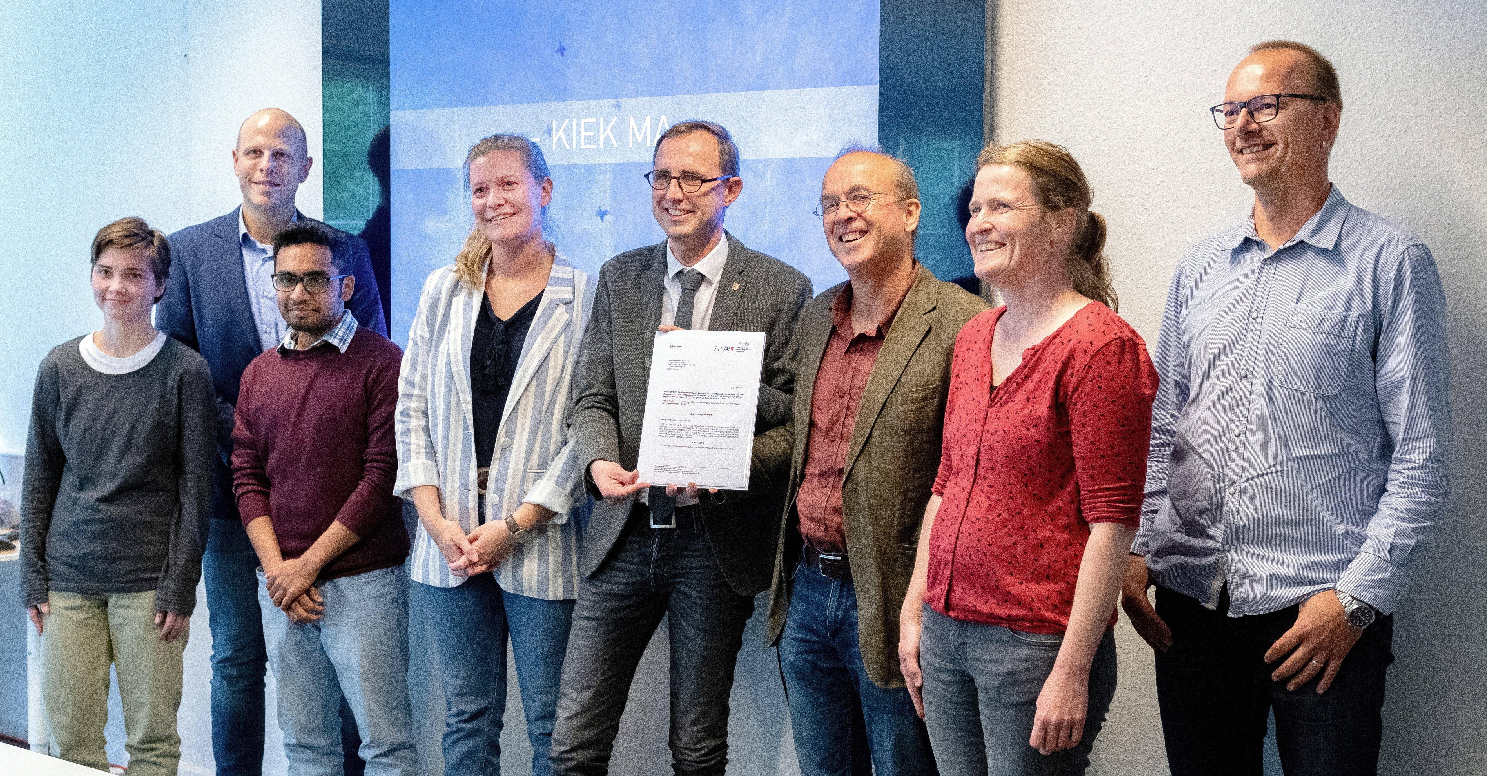 Eight people stand side by side in a room in front of a screen, the two people in the middle jointly hold a letter up to the camera.