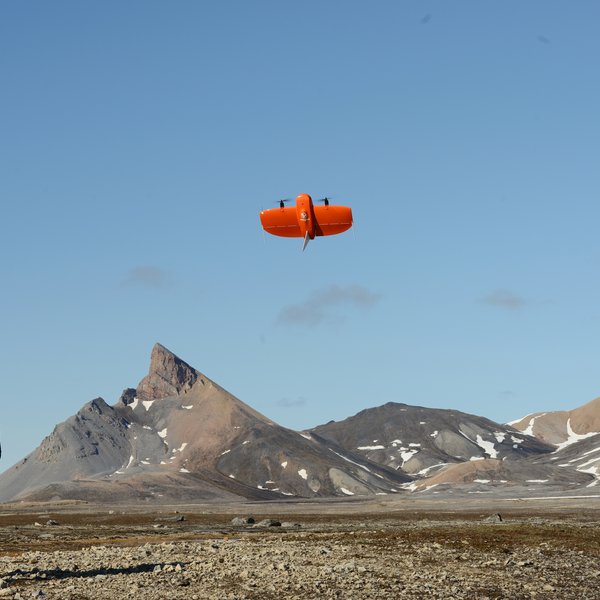 Ein Mann steht in einer weiten flachen Landschaft, vor ihm startet eine Drohne, im Hintergrund Berge.