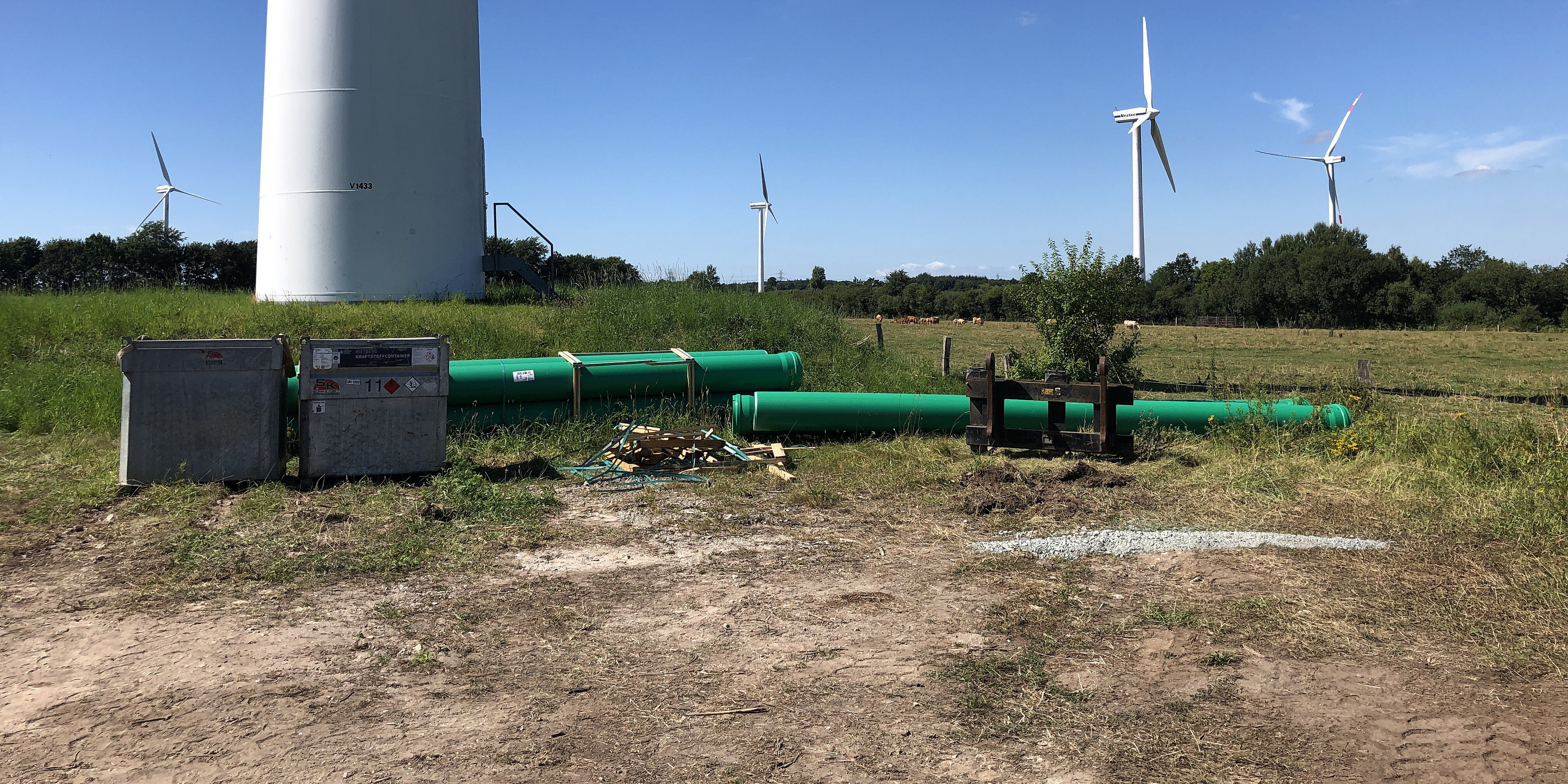 Windräder an Land, die auf Wiesen stehen. Im Vordergrund abgelegte Rohre und andere Baumaterialien. 
