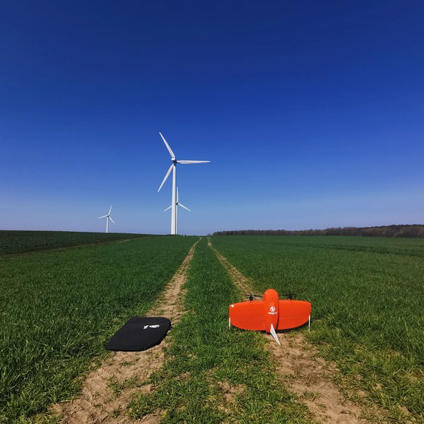 An orange drone stands on a field path in front of three wind turbines.