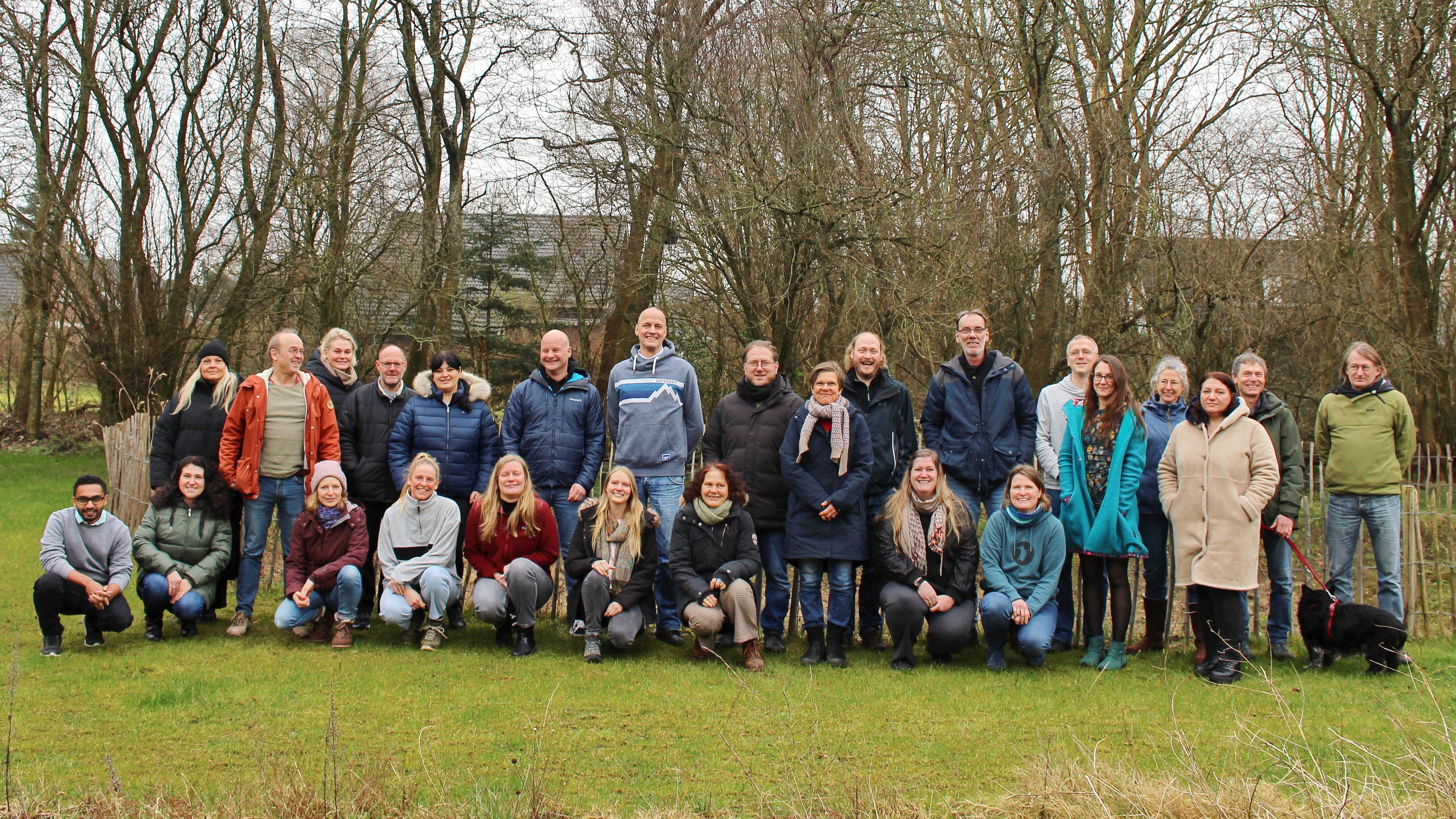 A group of people stand on a lawn for a group photo. Trees in the background.