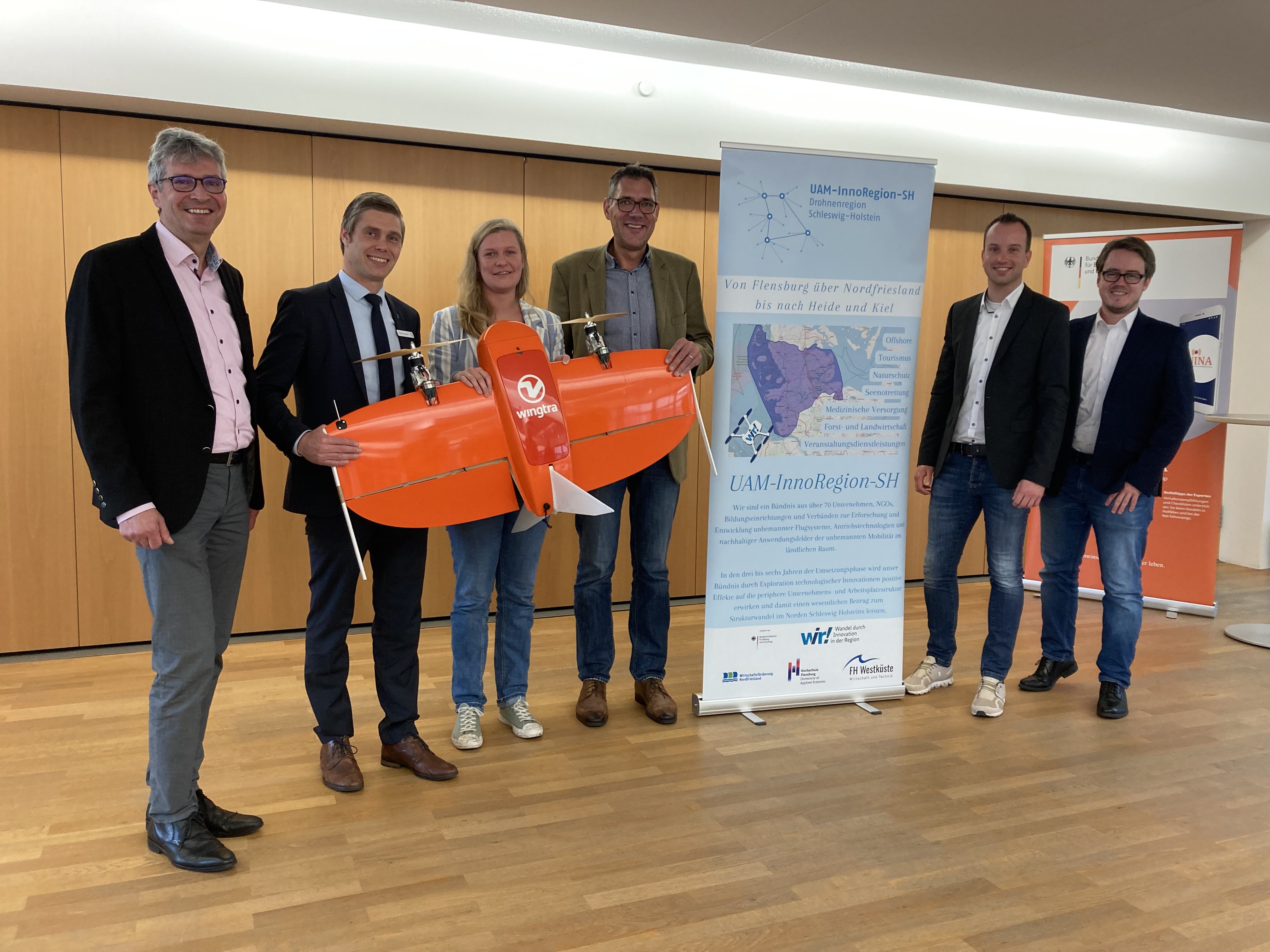 Six people stand next to each other, three of them holding a flight drone in their hands.