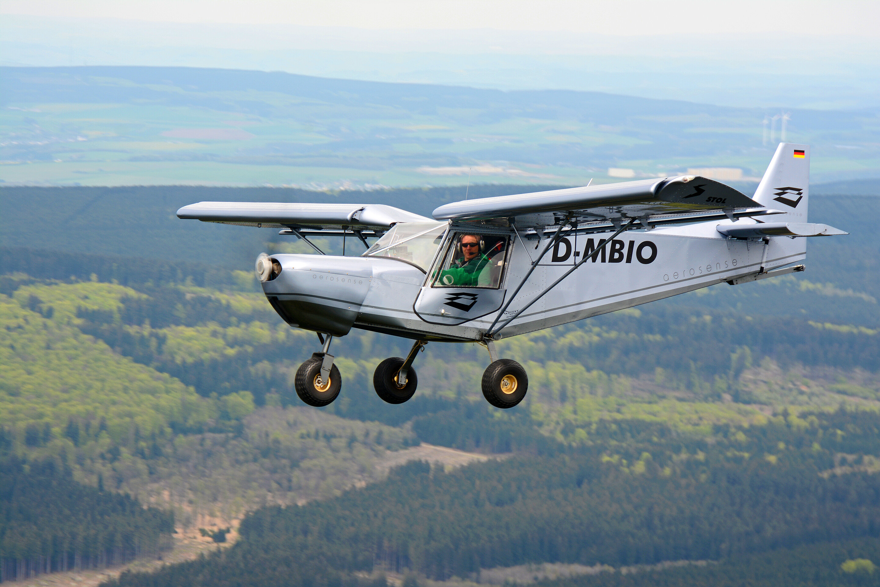 The silver light aircraft, used for aerial wildlife monitoring, flies over a wooded low-mountain landscape