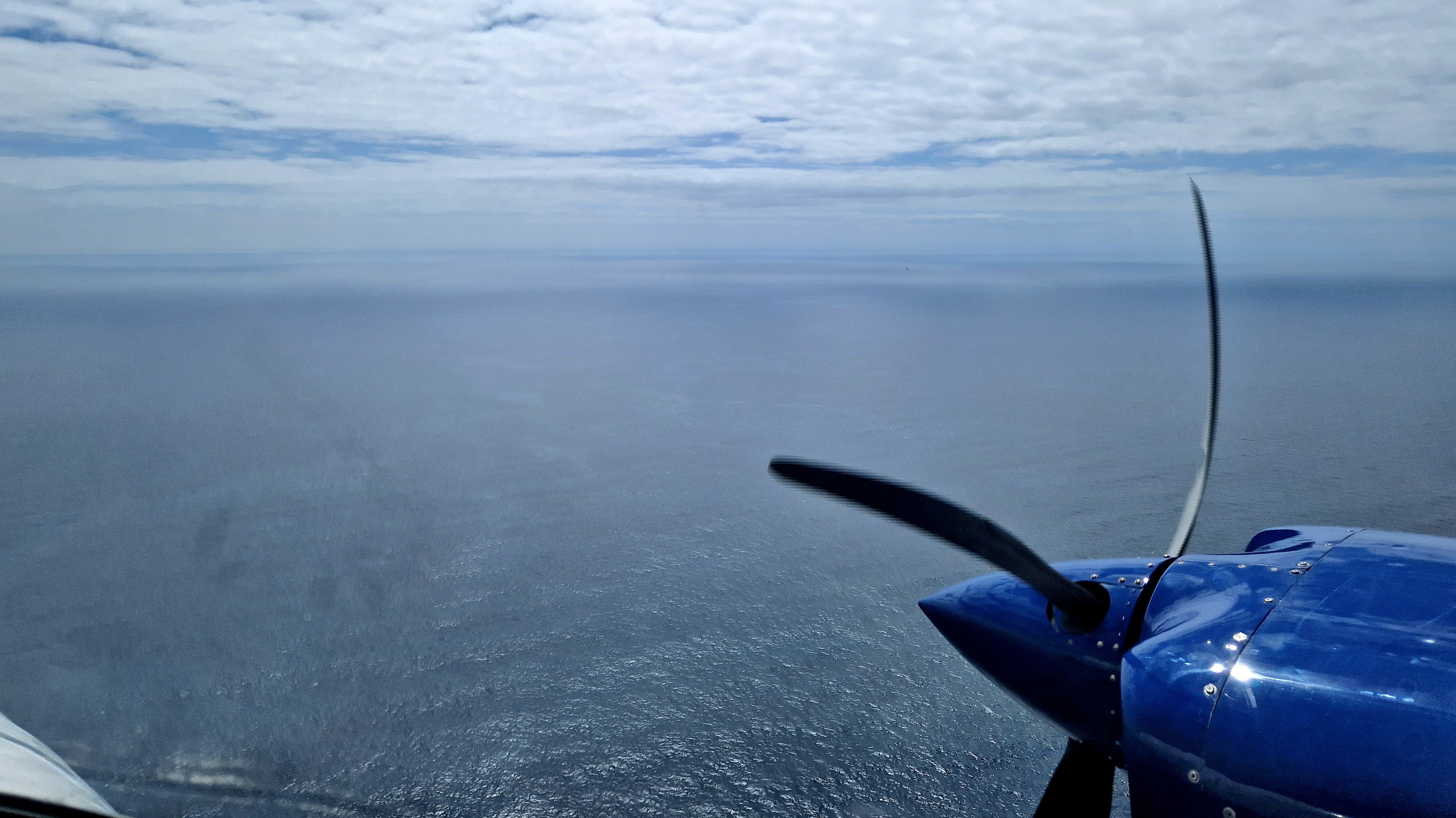 Blick aus einem Flugzeugfenster auf das Meer, vorne rechts im Bild der Propeller des Flugzeugs.