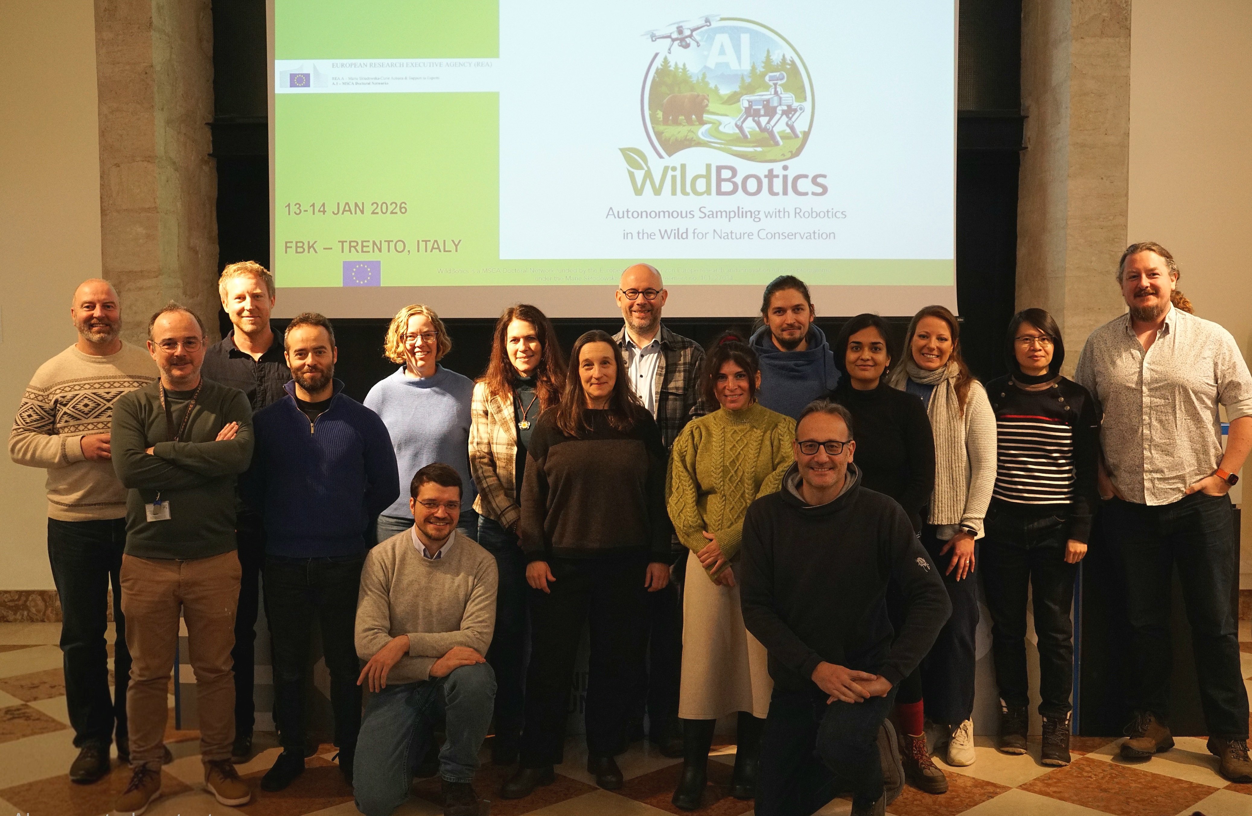 Seventeen people stand in front of a screen displaying the word ‘WildBotics,’ among other things, for a group photo.