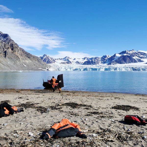 Zwei Männer auf dem Weg zu einem Boot am Strand einer Bucht auf Spitzbergen. Auf der anderen Seite der Bucht im Hintergrund ein Gletscher.
