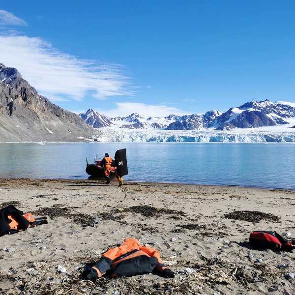 Two men on their way to a boat on the beach of a bay on Svalbard. On the other side of the bay, a glacier in the background.