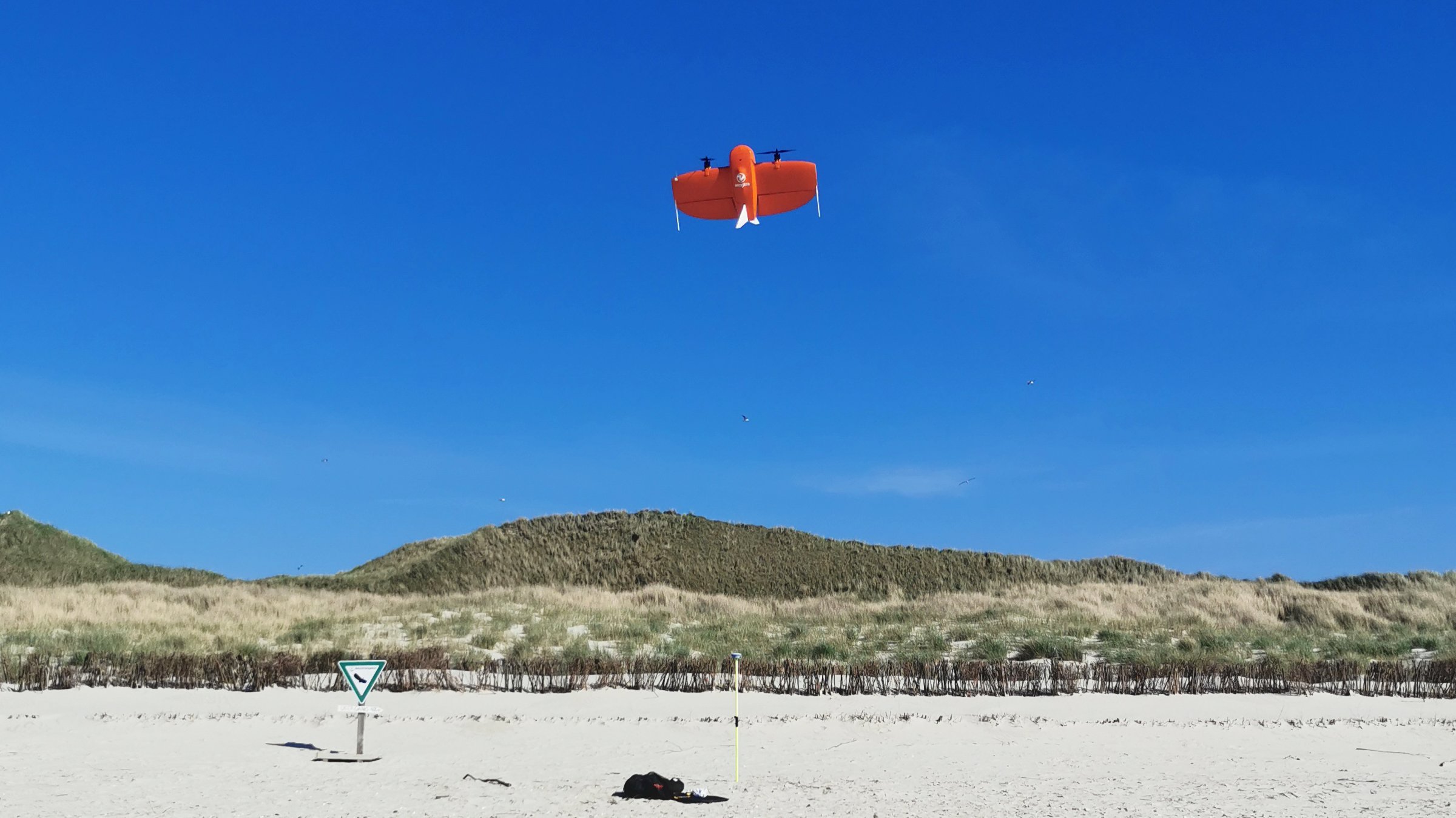 Eine Drohne startet vor blauem Himmel an einem Strand. Im Hintergrund Dünen.