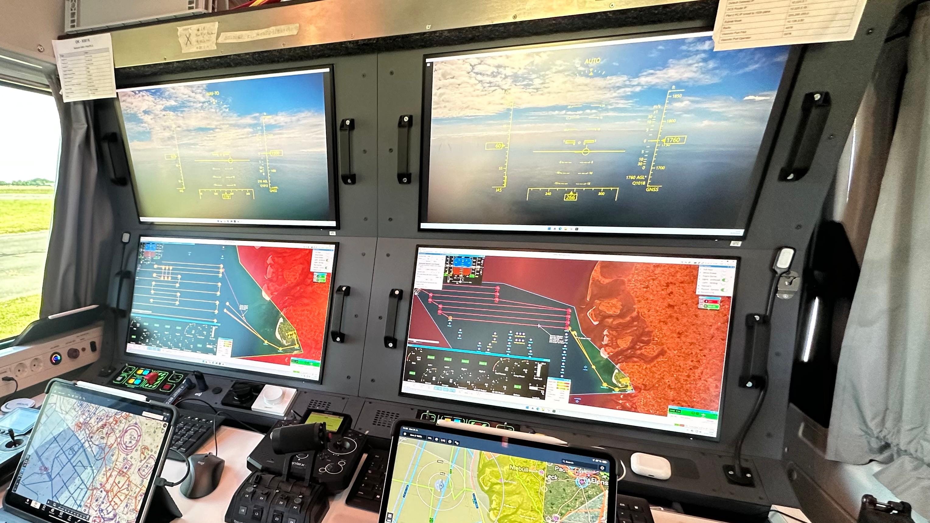 Blick in die Bodenstation (Ground Control Station) von der aus die Langstreckendrohnen von BioConsult SH geteuert werden. Vier große Bildschirme zeigen Kartenmaterial und den Blick von der Drohne nach vorne, wie sie in Echtzeit von einer Kamera übermittelt wird. Weiter im Vordergrund sind weitere Bedienungs- und Steuerungsgeräte, sowie zwei Laptops zu erkennen. 
