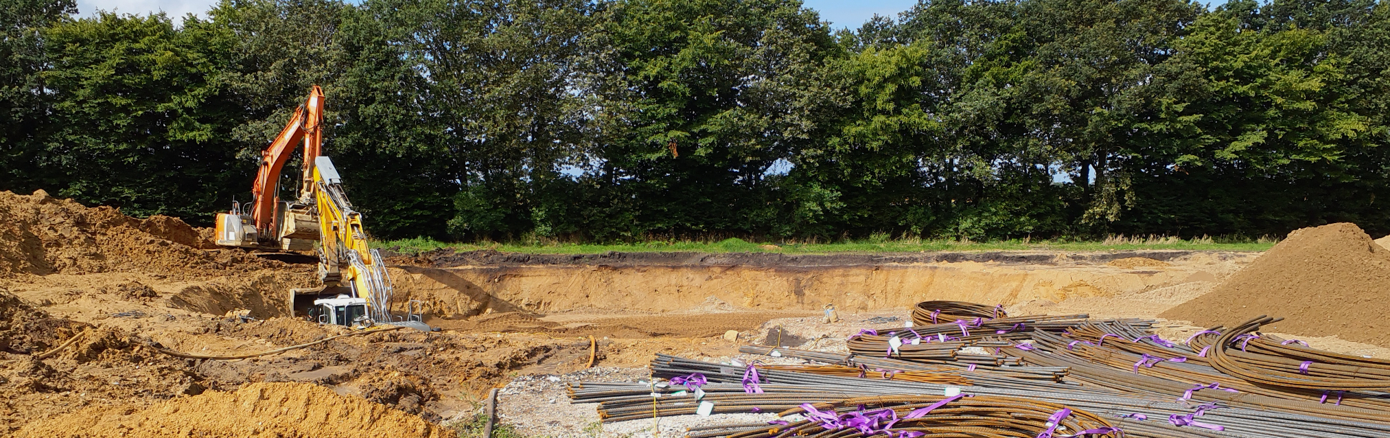 Baustelle mit zwei Baggern in einer großen Baugrube. Im Vordergrund liegen Bewehrungsstahlstäbe, im Hintergrund stehen Bäume.